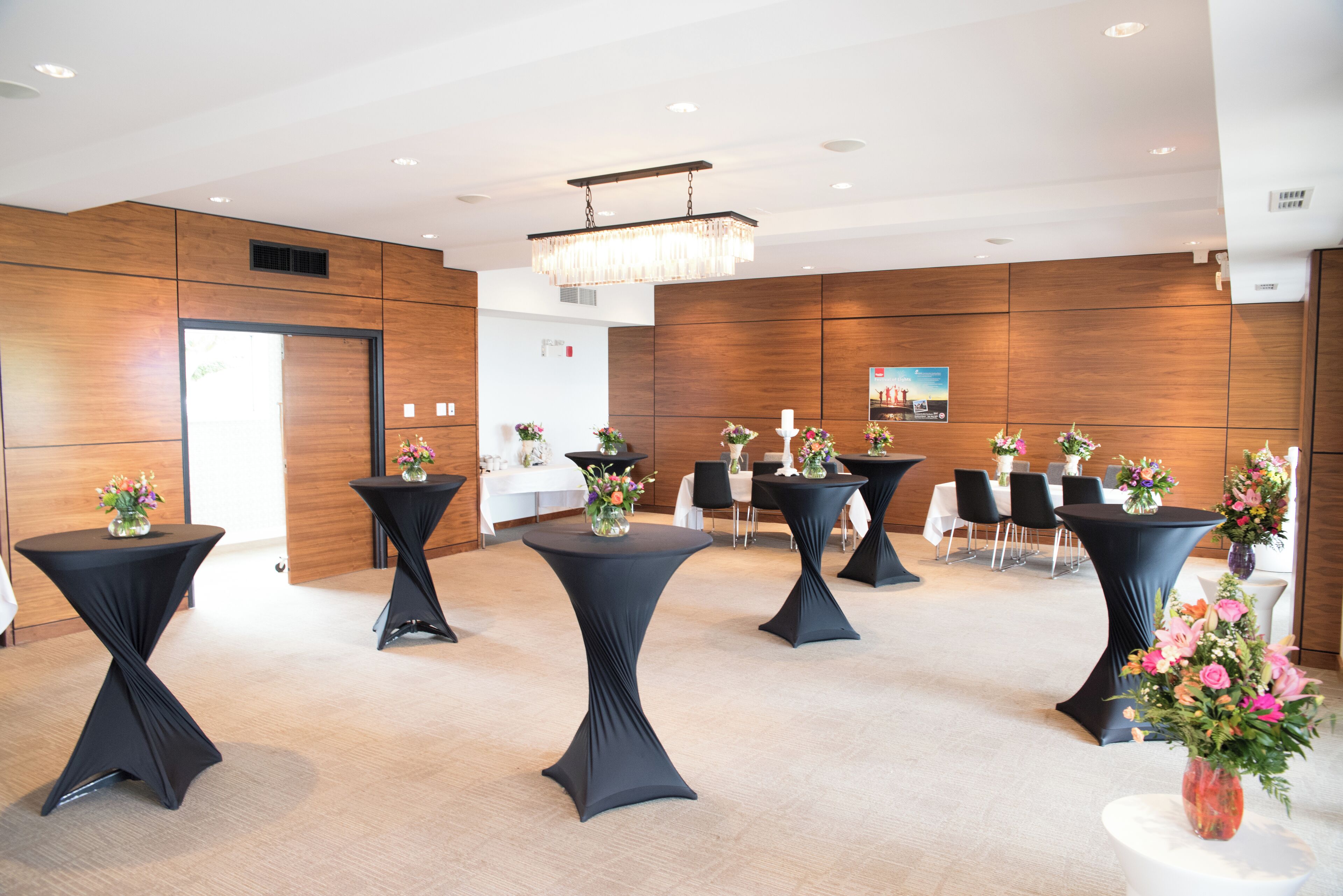A modern hotel lobby with wooden panel walls, black round tables with floral centerpieces, and a chandelier.