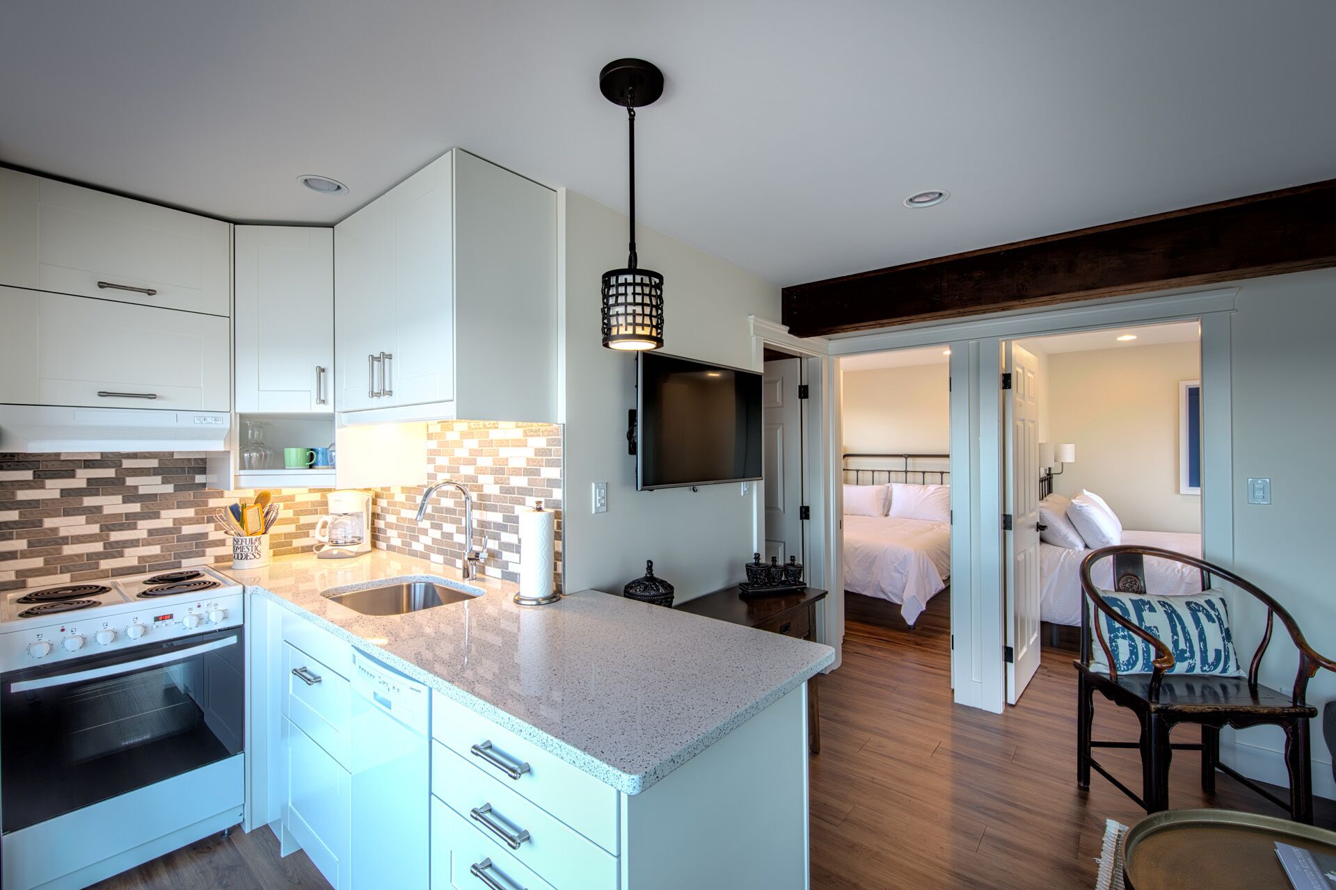 A modern kitchen with white cabinets, a built-in oven, and a large island with a sink. There is a dining area with a wooden table and chairs, and a bedroom with a bed and a nightstand.