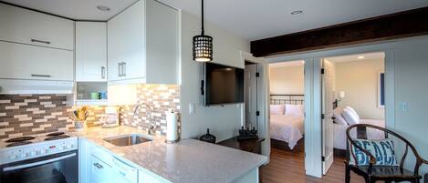A modern kitchen with white cabinets, a built-in oven, and a large island with a sink. There is a dining area with a wooden table and chairs, and a bedroom with a bed and a nightstand.