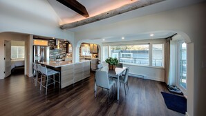 A modern kitchen with a central island, a dining area with a table and chairs, and large windows with blinds. - Qualicum Beach Ocean Suites  (Qualicum Beach)