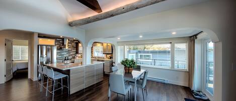 A modern kitchen with a central island, a dining area with a table and chairs, and large windows with blinds.