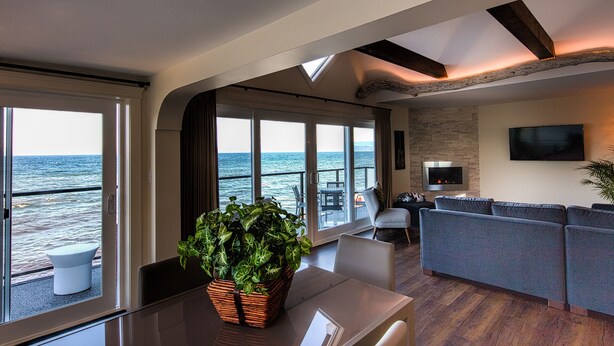 A modern living room with a large window overlooking the sea, a cozy seating area, and a flat-screen TV mounted on the wall. - Qualicum Beach Ocean Suites  (Qualicum Beach)