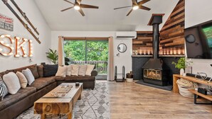A cozy living room with a brown leather sofa, wooden coffee table, and a fireplace. There are ceiling fans, a large window, and a sign that reads "SKI". - Prime Location on Camelback Mtn, Renovated w/ Movie & Game Room | The Bear's Den (Tannersville)