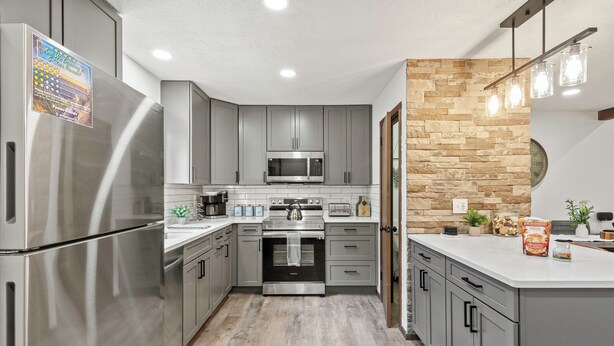 A modern kitchen with grey cabinets, stainless steel appliances, a stone accent wall, and pendant lighting. - Prime Location on Camelback Mtn, Renovated w/ Movie & Game Room | The Bear's Den (Tannersville)