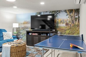 A living room with a flat-screen TV, a ping pong table, a blue sofa, and a wall mural of a forest scene. - Prime Location on Camelback Mtn, Renovated w/ Movie & Game Room | The Bear's Den (Tannersville)