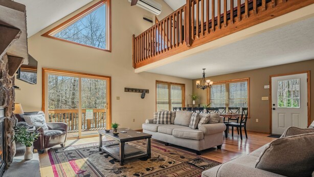A spacious living room with a high ceiling, large windows, a wooden staircase, and a dining area.