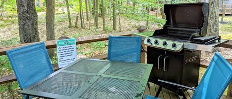 An outdoor grill area with a table and chairs, surrounded by trees.