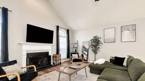 Ein Wohnzimmer mit Kamin, einem flachbildfernseher darüber, einem grünen Sofa, einem Holztisch und einem schwarzen Sessel.