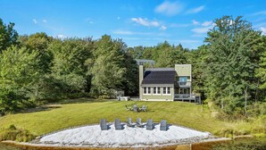 A modern house with a large deck, surrounded by a pond and dense trees. - Strawberry Shores by AvantStay | Lakefront Private Beach (Long Pond)