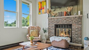 A living room with a brick fireplace, a large window, a comfortable armchair, and a coffee table. - Strawberry Shores by AvantStay | Lakefront Private Beach (Long Pond)