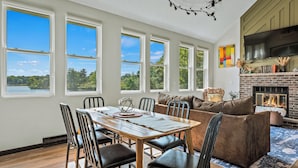 A dining area with a wooden table and chairs, a fireplace, and a large window with a view of trees and a lake. - Strawberry Shores by AvantStay | Lakefront Private Beach (Long Pond)