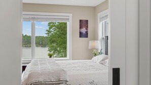 A bedroom with a large window, a bed with a white quilt, a bedside table with a lamp, and a wall-mounted artwork. - Newly Built Lakefront w/ Deck & Views | Lakeside Serenity by AvantStay (White Haven)