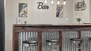 A bar area with a wooden counter, three bar stools, a 'Bar' sign, and two wall-mounted light fixtures. - Ridge Retreat by AvantStay | Game Room, Covered Porch, & Fire Pit! (Blakeslee)