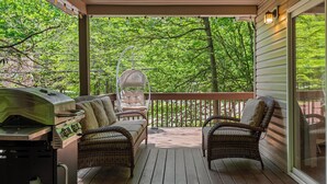 Um deck coberto com churrasqueira, cadeiras de vime e uma porta de vidro que dá para uma área verde exuberante.