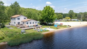 A lakeside cabin with a deck, surrounded by greenery and a sandy beach. - The Lake Life by AvantStay | Lakefront + View + Hot Tub (Pocono Lake)