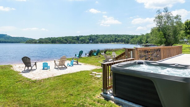A lakeside deck with Adirondack chairs, a hot tub, and a wooden deck overlooking a lake. - The Lake Life by AvantStay | Lakefront + View + Hot Tub (Pocono Lake)