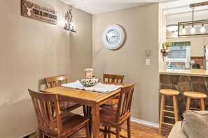 Un comedor con una mesa de madera, cuatro sillas, un reloj en la pared y una cocina a lo lejos.