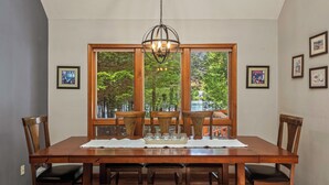 Un coin repas comprenant une table en bois, six chaises, un lustre et de grandes fenêtres donnant sur la forêt.