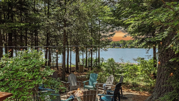 Une zone boisée où sont disposées des chaises Adirondack autour d’un foyer, avec vue sur un lac.