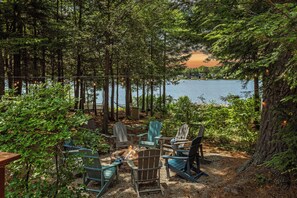 A forested area with Adirondack chairs arranged around a fire pit, overlooking a lake.