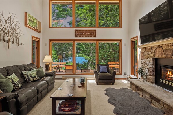 A spacious living room with a leather sofa, a coffee table, and a fireplace. Large windows offer a view of the outdoors.