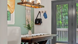A dining area with a wooden table, a white chair, a hanging light fixture, a plant, and a view of a deck through a glass door.