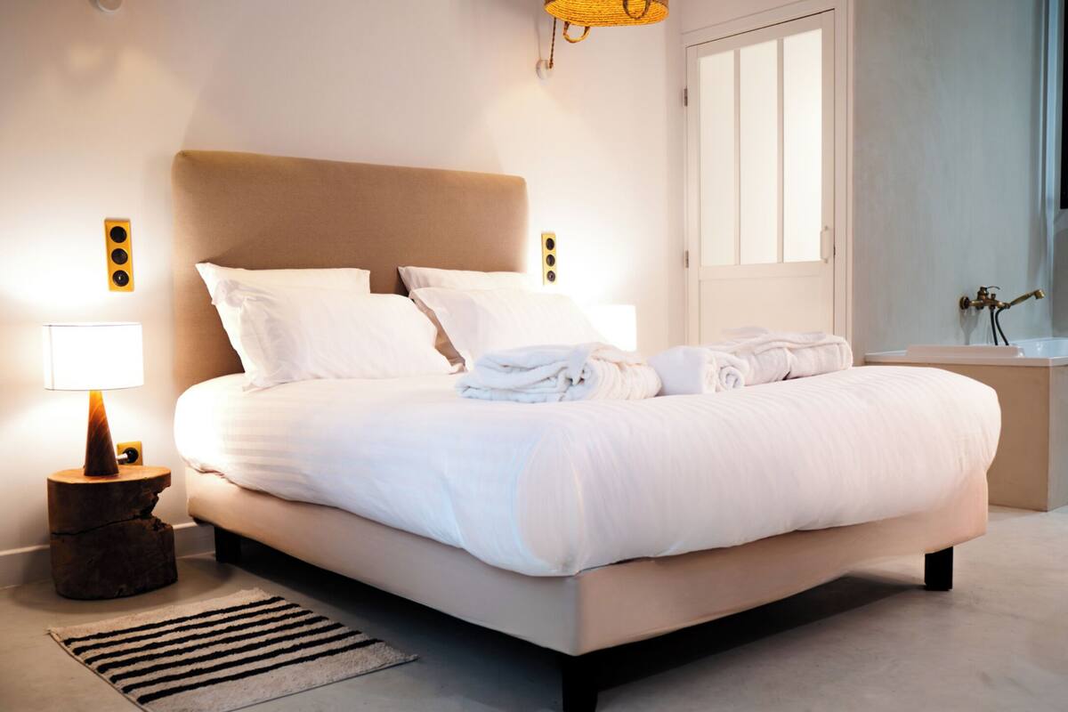Una cama bien hecha con sábanas blancas, una lámpara de noche y una mesita de noche de madera con una lámpara.