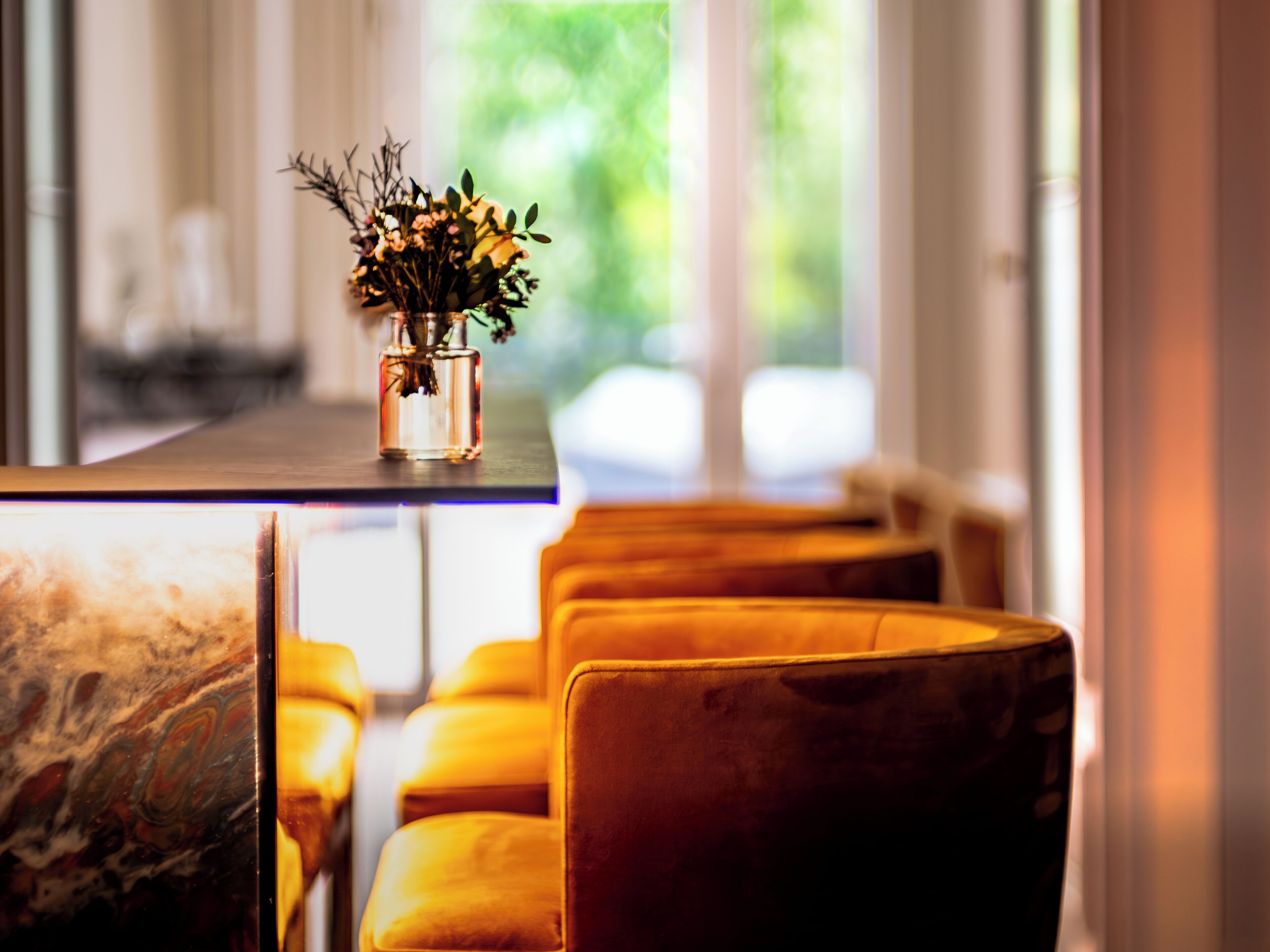 A modern interior with a long table and yellow chairs, a vase with flowers, and a view of greenery outside.