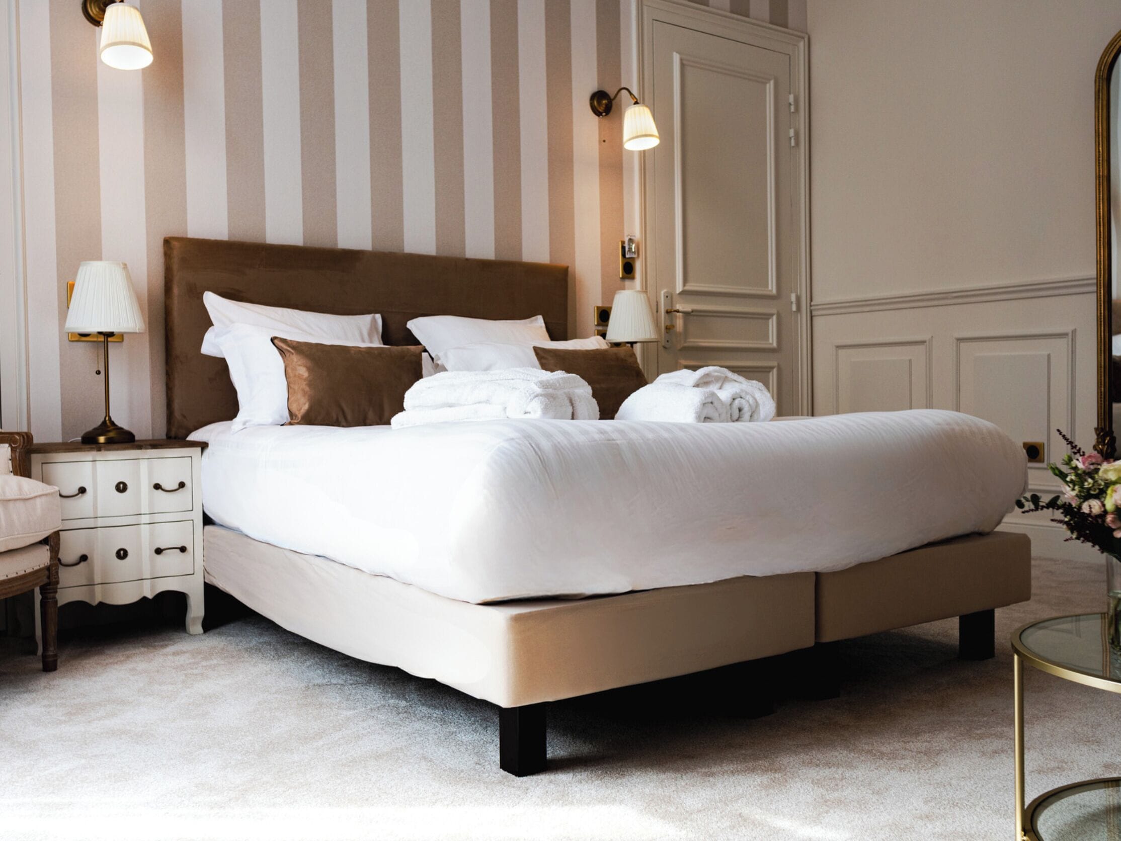 A neatly made bed with white linens and brown pillows in a hotel room with striped wallpaper and a large mirror.