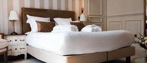 A neatly made bed with white linens and brown pillows in a hotel room with striped wallpaper and a large mirror.
