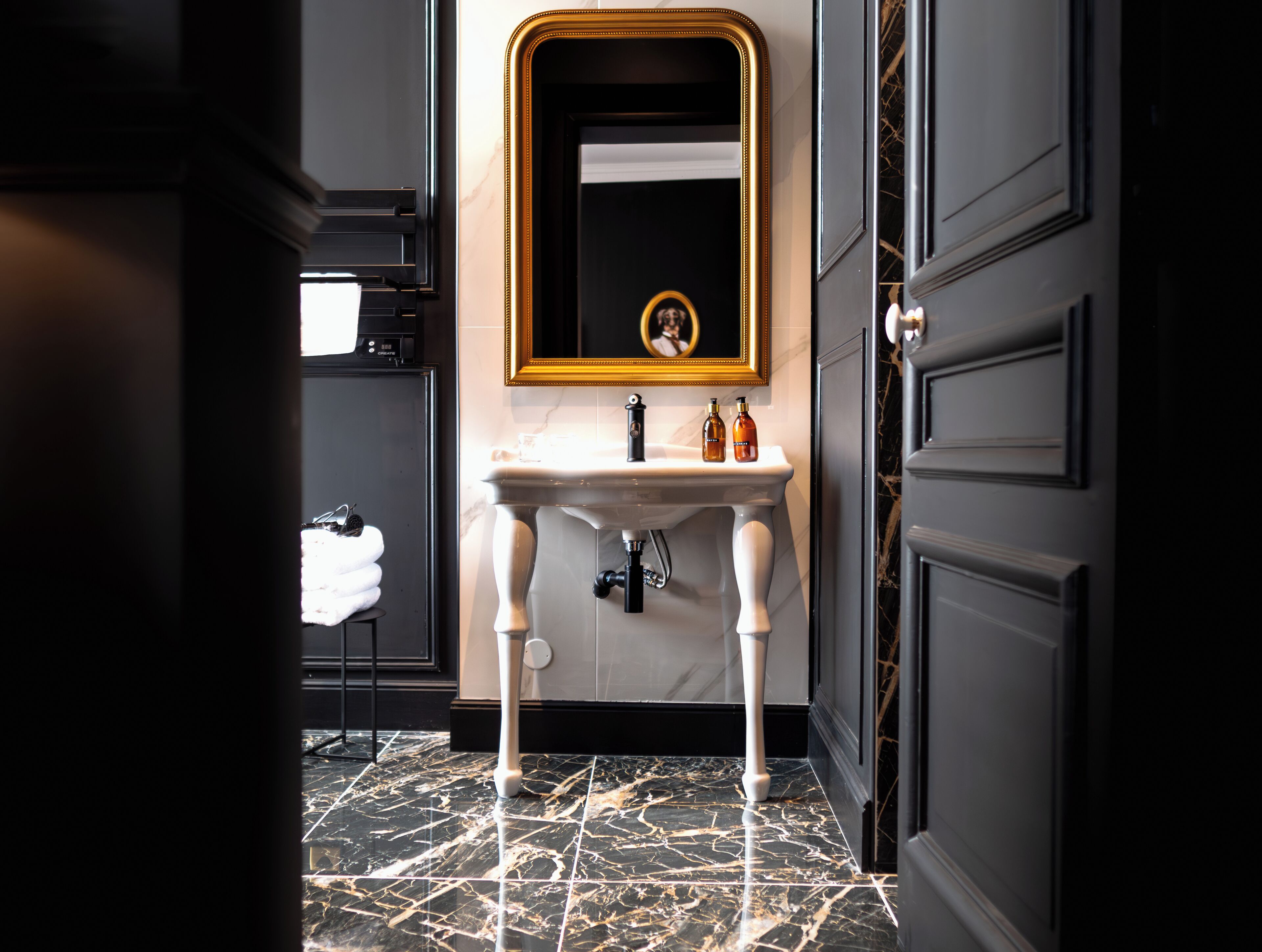 Een badkamer met een marmeren vloer, een wastafel op voet en een spiegel met gouden lijst.