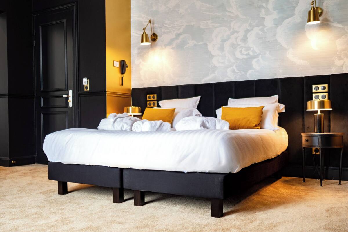 Una cama grande con ropa de cama blanca y dos almohadas amarillas, un cabecero oscuro y una pared con un diseño de nubes.