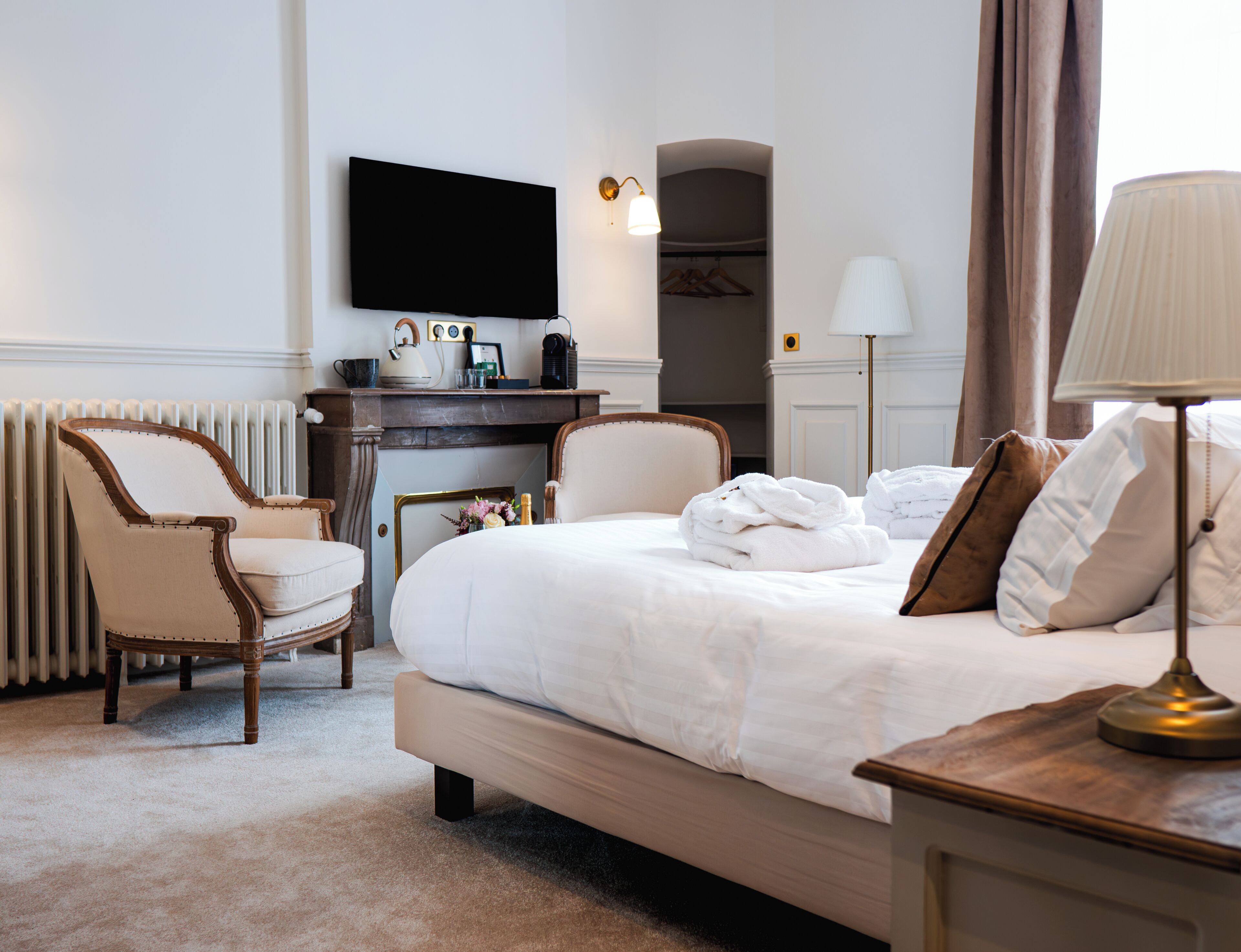 A hotel room with a bed, a desk with a flat-screen TV, a chair, a lamp, and a radiator.