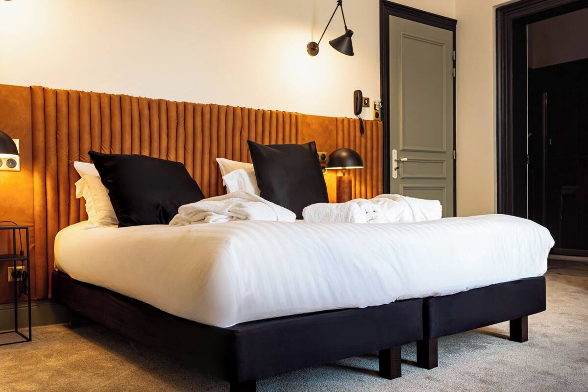 Una cama bien tendida con sábanas blancas y almohadas negras, cabecera de madera, lámpara de noche y un teléfono en la pared.