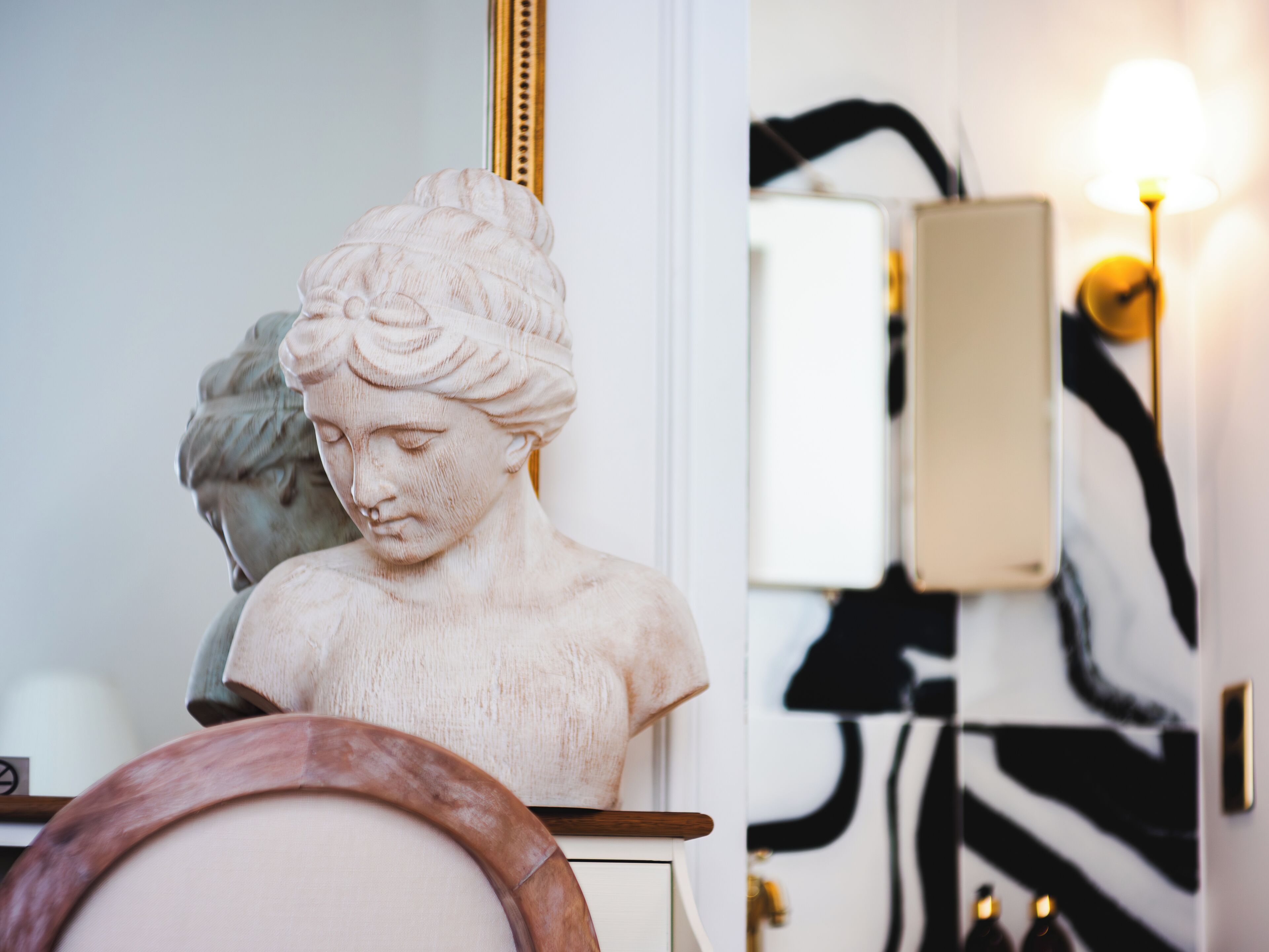 Un buste sculpté posé sur une table en bois, un miroir et un mur à rayures zébrées.
