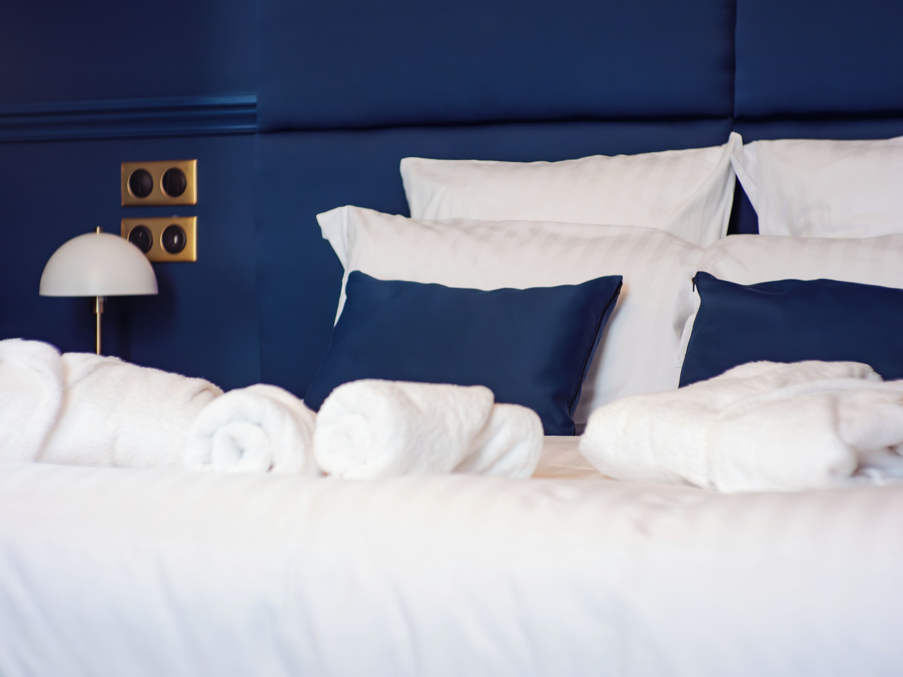Een opgemaakt bed met witte lakens en blauwe kussens, een gouden speaker en een witte lamp op een blauwe muur.