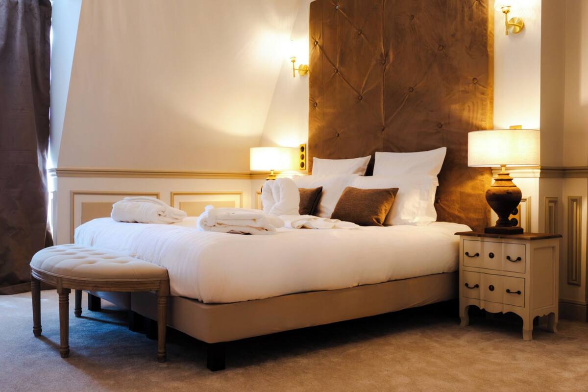 Una cama bien hecha con almohadas y una colcha, una mesita de noche de madera con cajones y una lámpara.