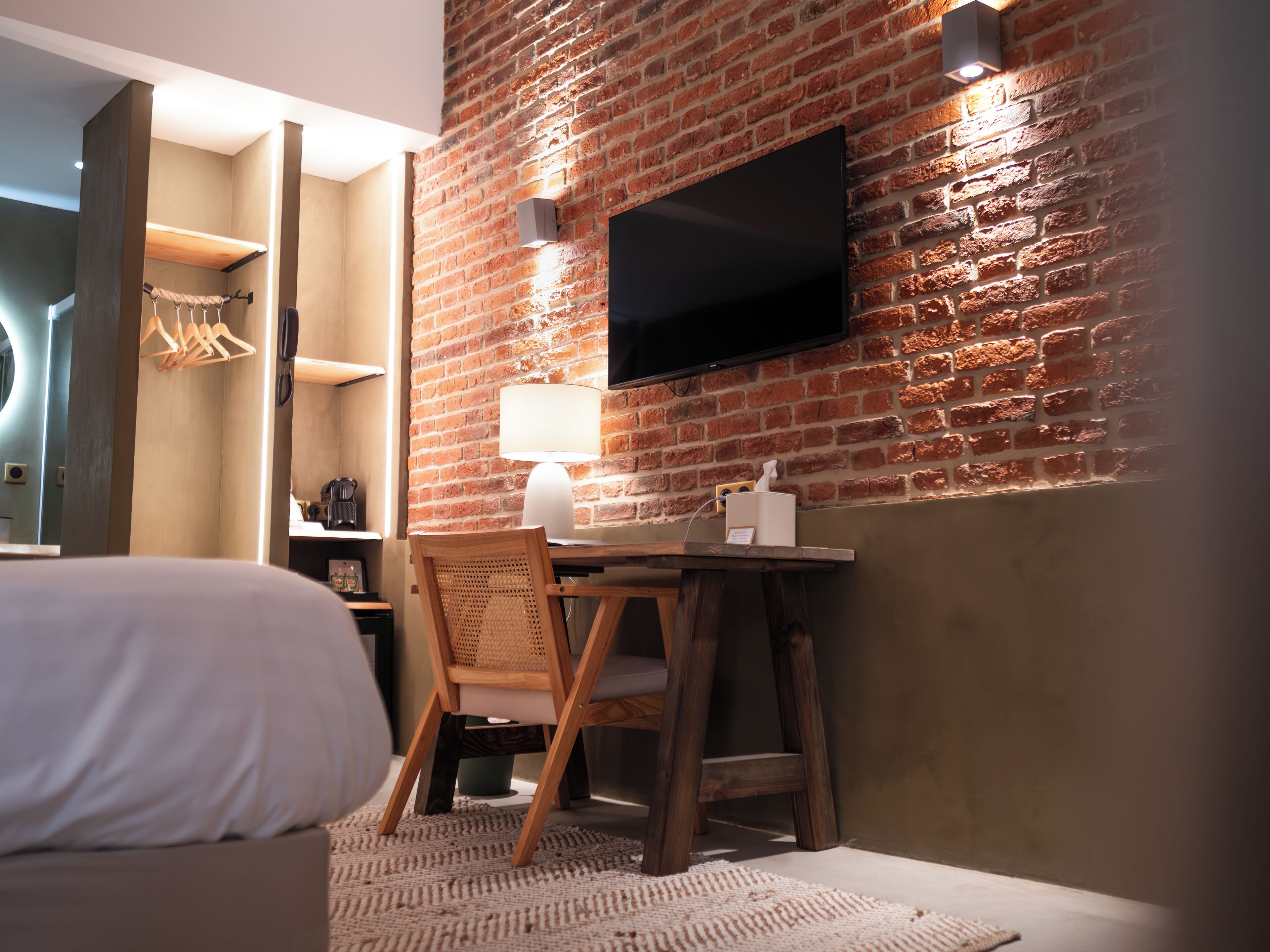 A hotel room with a brick wall, a flat-screen TV mounted above a wooden table, a chair, and a bed with a white pillow.