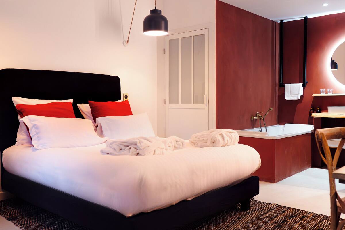Una cama bien hecha con almohadas blancas y rojas, un cabecero negro y una mesita de noche con una lámpara. Hay un baño con un lavabo, espejo y toallero.