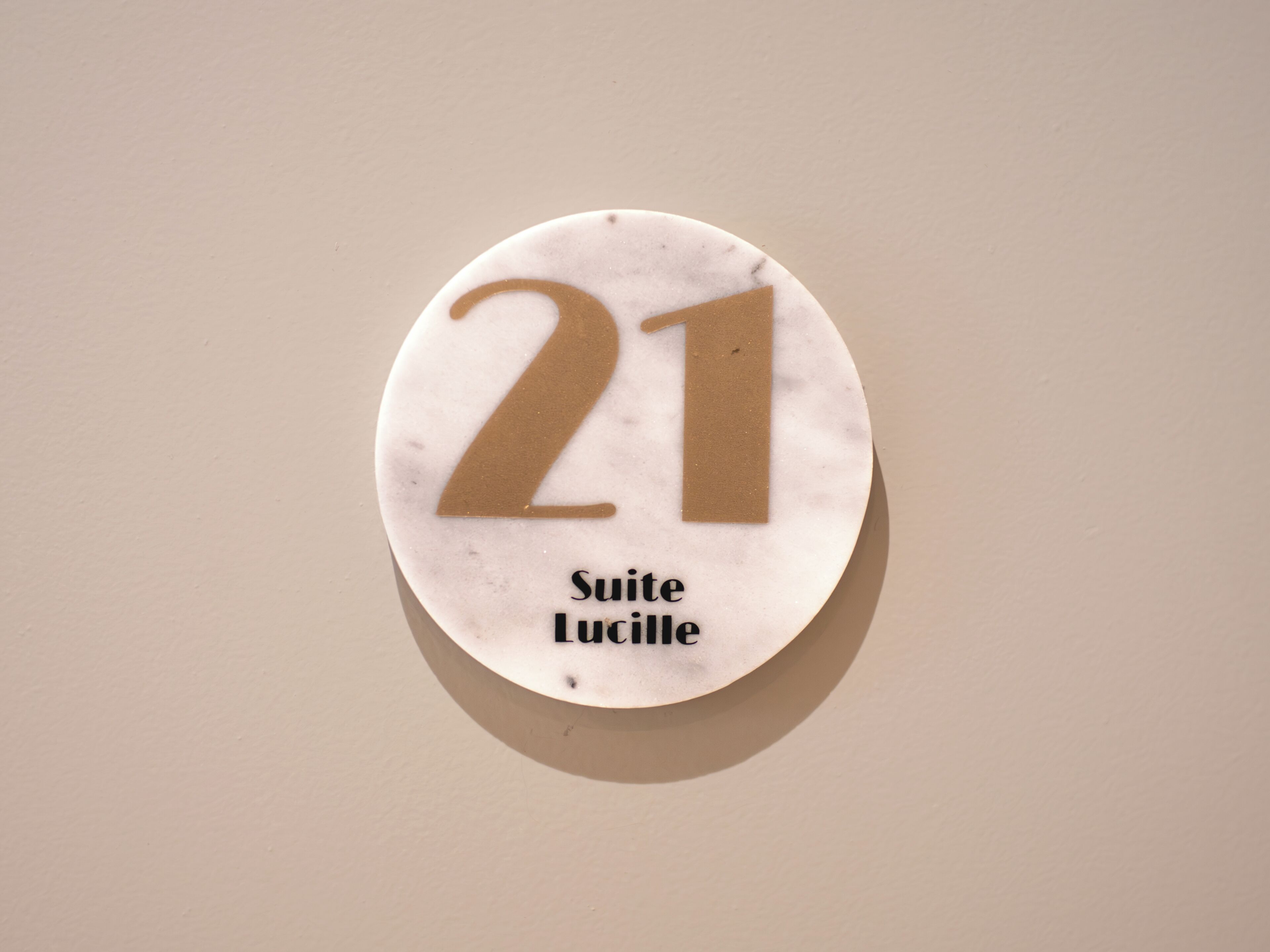 Un panneau circulaire portant le numéro 21 et l’inscription « Suite Lucille » sur un fond uni.