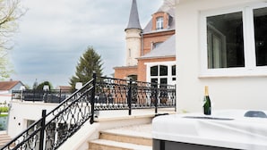 A staircase with ornate black railings leading to a white building with a large window. In the background, there is a brick building with a tall tower. - Hotel Château Héloïse (Mérélessart)