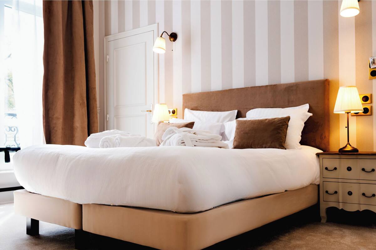 Una cama bien hecha con sábanas blancas y almohadas marrones, una mesita de noche con una lámpara, y una pared a rayas al fondo.