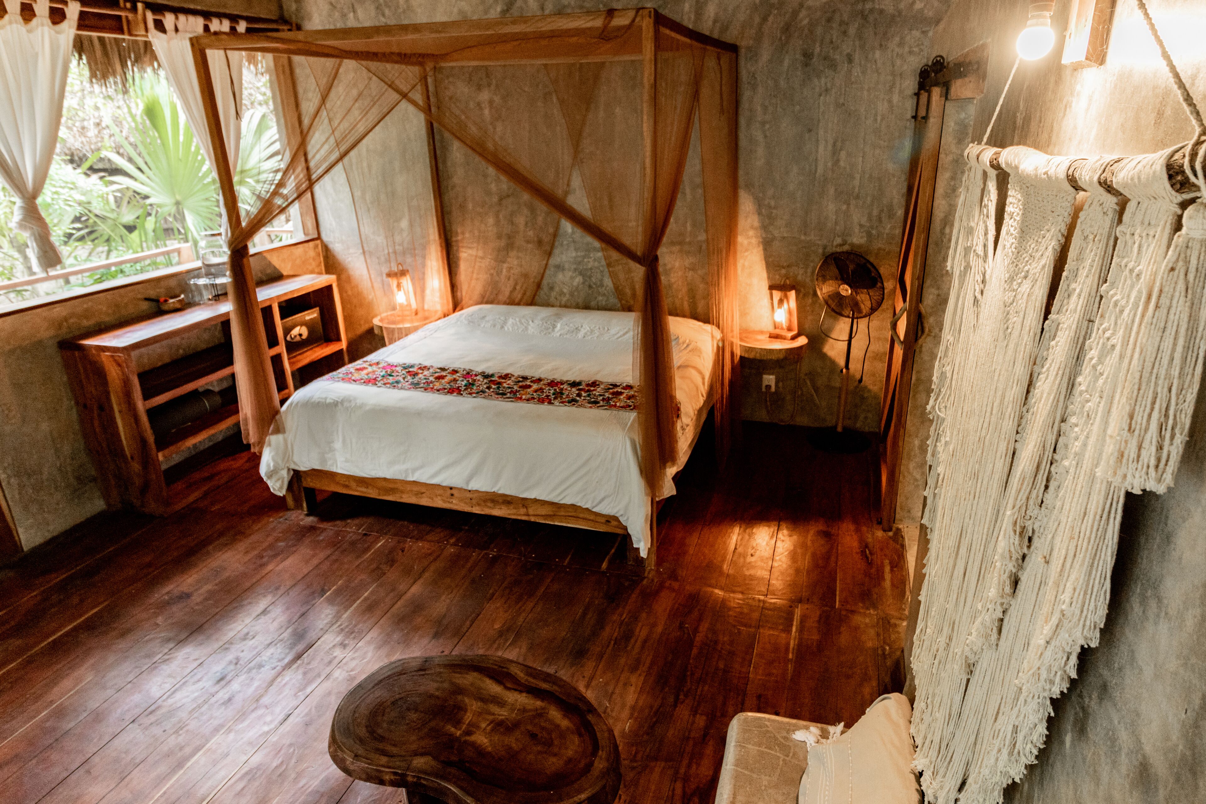 A room with a four-poster bed, wooden furniture, and a view of greenery outside.