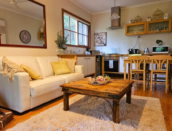 Una sala de estar con un sofá blanco, una mesa de centro de madera con un frutero, una zona de cocina con armarios de madera, un microondas y una mesa de comedor con sillas.