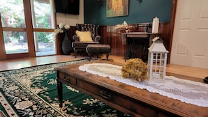A living room with a large green patterned rug, a wooden coffee table, a television mounted on the wall, a fireplace, and a large window with a view of greenery. - Memorable Gatherings at FernglenForestRetreat (Mount Dandenong)