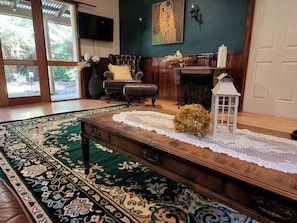 A living room with a large green patterned rug, a wooden coffee table, a television mounted on the wall, a fireplace, and a large window with a view of greenery.