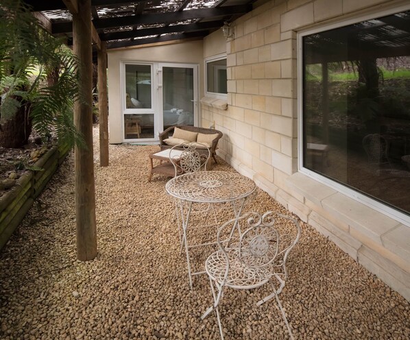 Un patio couvert aménagé avec un canapé en osier et deux tables en métal, le tout entouré de galets.