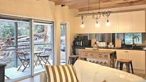 A modern living room with a white sofa, striped pillows, and a glass coffee table. There is a kitchen area with white cabinets and a dining space with a table and chairs. - Memorable Gatherings at FernglenForestRetreat (Mount Dandenong)