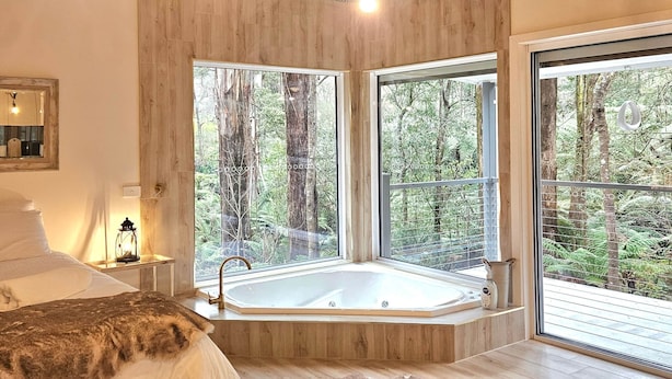 A modern bedroom with a large bed, a wooden floor, a fur rug, a glass bathtub, and large windows with a forest view. - Memorable Gatherings at FernglenForestRetreat (Mount Dandenong)