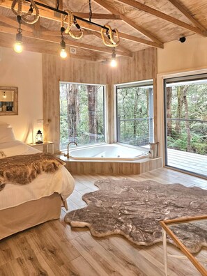Une chambre moderne avec un grand lit, un plancher en bois, un tapis en fourrure, une baignoire en verre et de grandes fenêtres donnant sur une forêt.